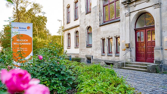 Ein historisches Steinhaus mit hohen Fenstern und einer roten Tür, umgeben von blühenden rosa Rosen. Neben dem Weg, der zum Eingang führt, steht ein Schild auf Deutsch mit der Aufschrift „Herzlich Willkommen!“.