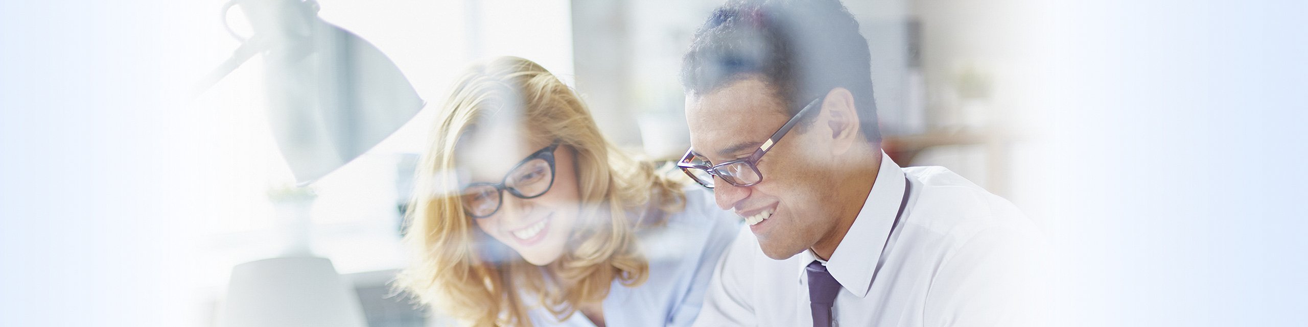 Zwei lächelnde Personen, ein Mann mit Brille und Krawatte und eine Frau mit Brille, die gemeinsam an einem Schreibtisch im Büro arbeiten