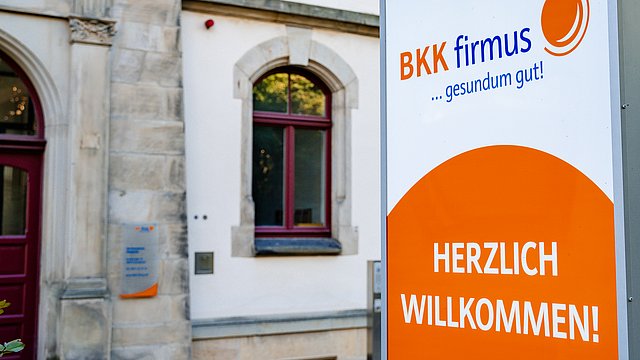 Ein Schild der Krankenkasse „BKK firmus“ heißt Besucher vor einem Gebäude mit Bogenfenstern willkommen.