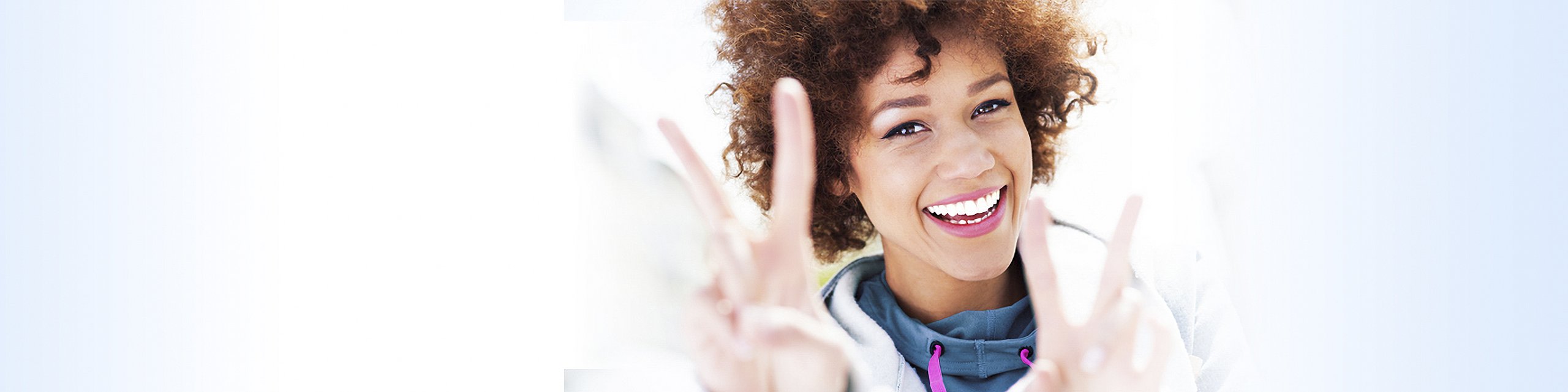 Lächelnde Frau mit lockigen Haaren, die mit beiden Händen das Friedenszeichen zeigt, vor hellem Hintergrund