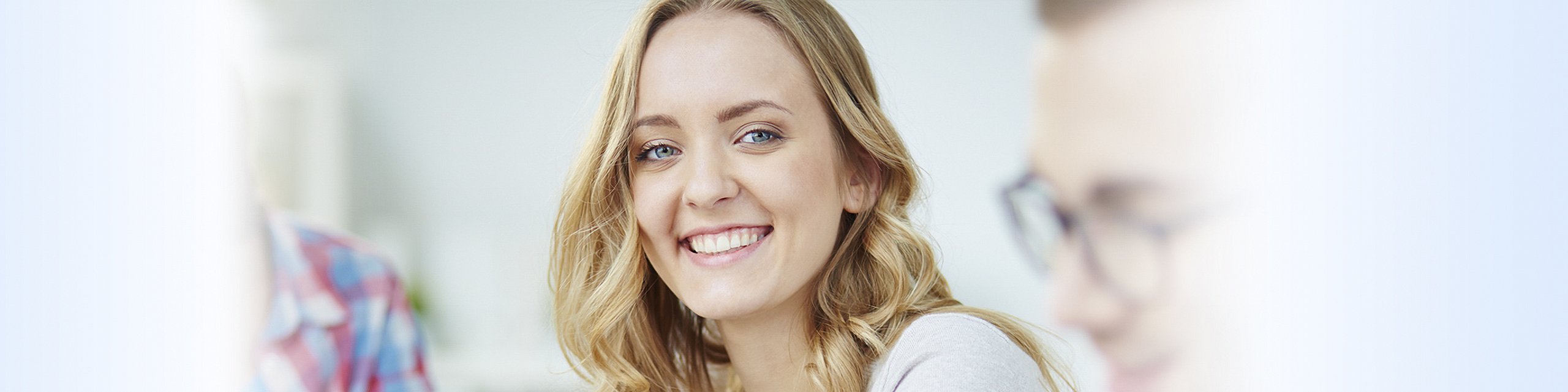 Lächelnde Frau mit langen blonden Haaren in einem Büro, im Hintergrund Kollegen und Computer