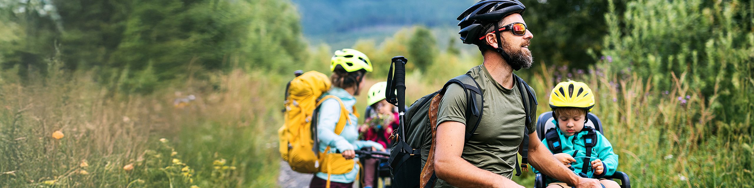 Eine junge Familie in sportlicher Kleidung steht mit Fahrrädern auf einer Wiese, die beiden Kinder sind auf den Fahrrädern im Kindersitz.