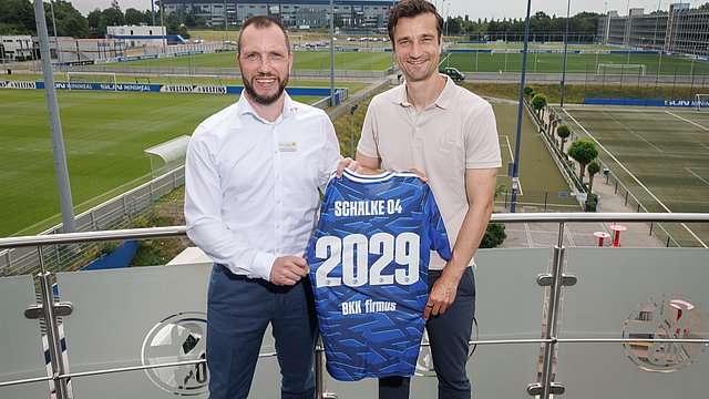 Zwei Männer stehen auf einem Balkon einer Fußballtrainingsanlage, lächeln und halten ein blaues Schalke-04-Trikot in den Händen, auf dessen Rücken die Zahl 2029 aufgedruckt ist. Im Hintergrund sind das Fußballfeld und die Tribünen zu sehen.