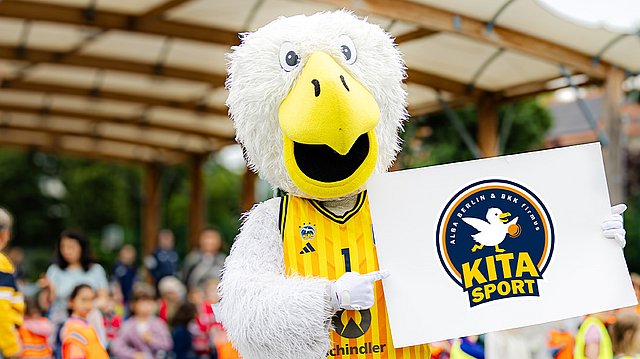 Das Maskottchen von ALBA Berlin hält bei einer Freiluftveranstaltung mit Kindern das Logo für den Kita-Sport hoch.