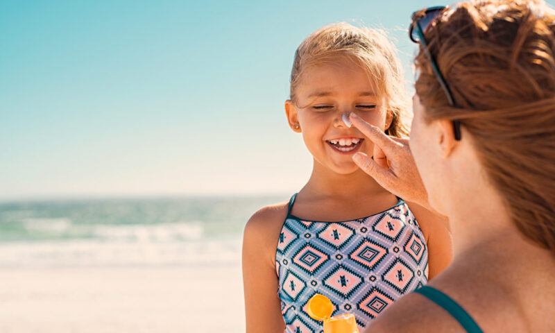 Mutter cremt ihre Tochter am Strand mit Sonnencreme ein.