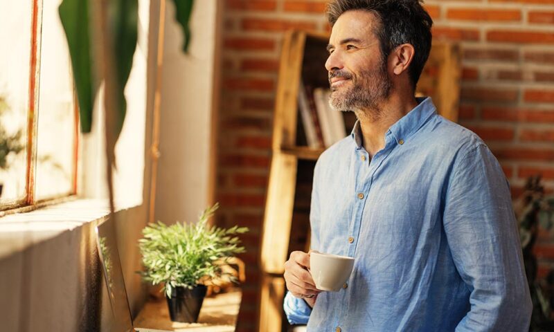 Lächelnder Mann mittleren Alters sieht aus dem Fenster hinaus und hält eine Tasse Kaffee in der Hand.